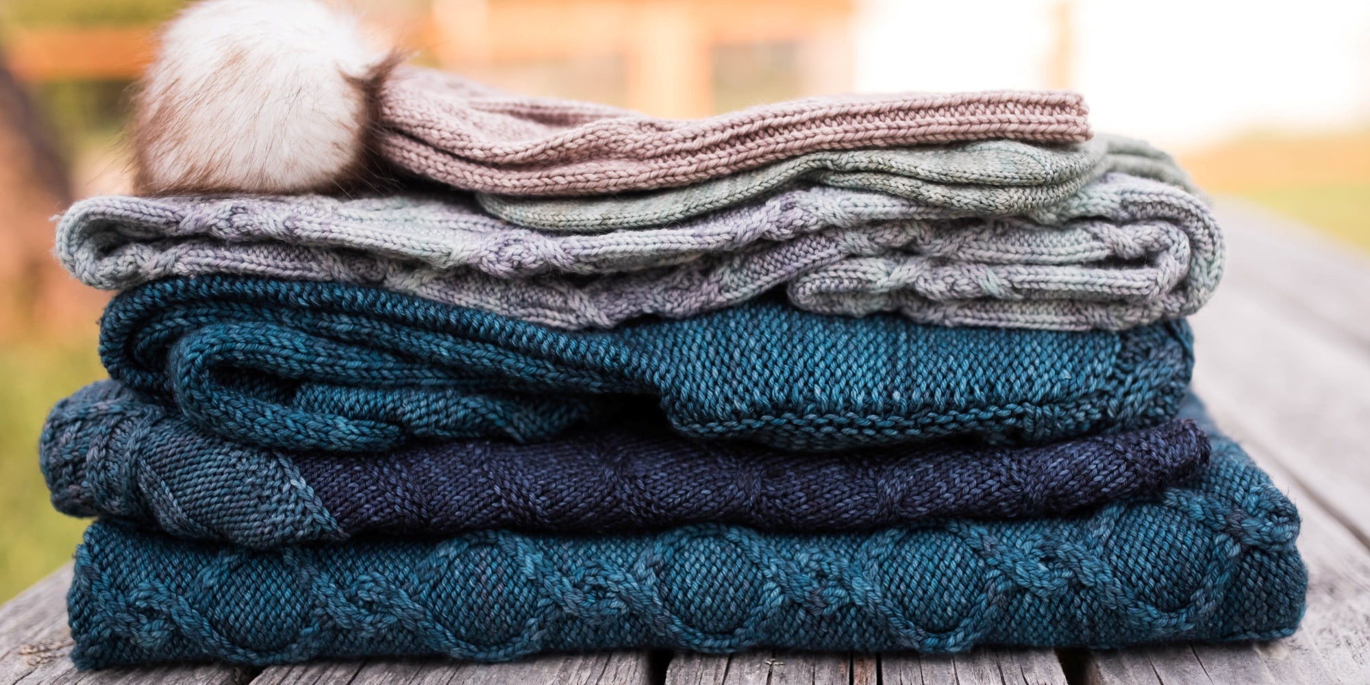 five knitted items, all cabled, in various shades of blue and beige, are folded in a squishy stack